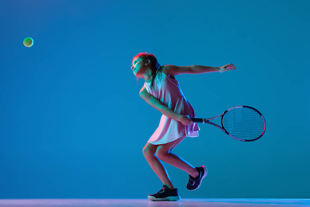 Studio shot of little girl, beginner tennis player training isolated on blue studio background in neon light. Sport, study, childhood concept - Foto, Imagem