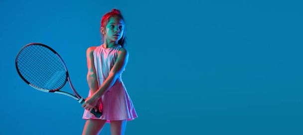 Studio shot of little girl, beginner tennis player training isolated on blue studio background in neon light. Sport, study, childhood concept - Foto, afbeelding