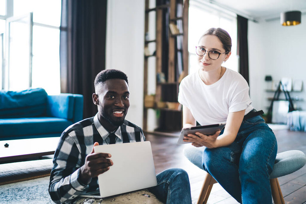 Pleasant woman and content black man in casual clothes sitting in creative workspace with laptop and tablet and looking at camera while studying together - Zdjęcie, obraz
