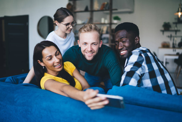 Positive woman in bright t shirt taking selfie on mobile phone while sitting on comfortable couch near multiracial colleagues in living room - Photo, Image