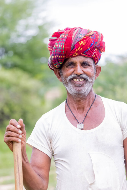 Jawai, Rajastán, India - septiembre de 2021: Retrato de un anciano del grupo étnico Rabari con un tocado nacional y vestimenta blanca tradicional y turbante rojo en el campo de Jawai. - Foto, Imagen
