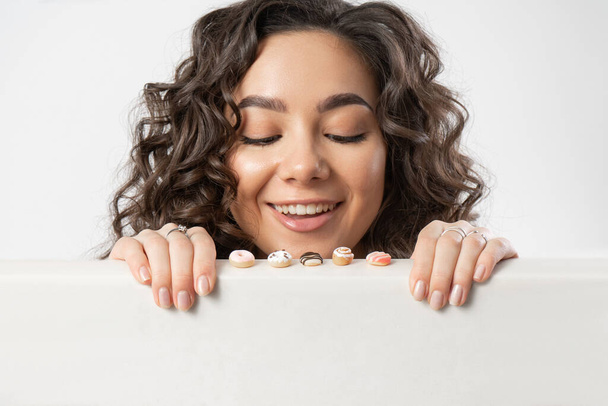Een mooie jonge vrouw met krullend lang donker haar en bruine ogen kijkt verrast en geïnteresseerd in miniatuur zoete donuts die ze willen eten - Foto, afbeelding