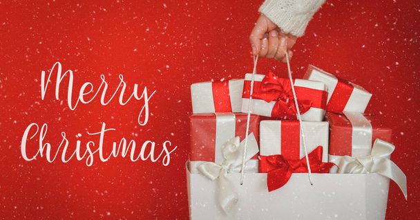 Merry christmas text. Female wearing white knitted sweater holding a shopping bag full of presents over isolated red background. Bunch of gifts in hands of a woman. Close up, copy space for text. - Photo, Image