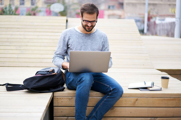 Positive male in casual jumper sitting on podium in street with backpack while working on computer during remote work in daylight - Fotó, kép