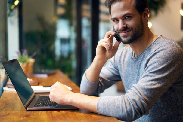 Crop side view of bearded male entrepreneur wearing casual clothing talking on mobile phone while sitting at wooden table with laptop - Foto, Imagen