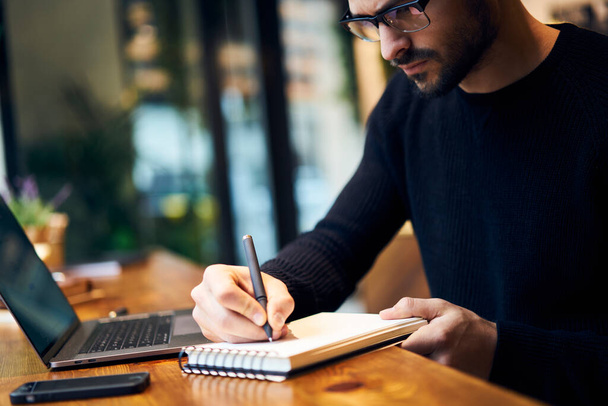 Crop side view of serious concentrated male freelancer in casual outfit and eyeglasses writing in notepad while sitting at wooden table with laptop and smartphone - Foto, afbeelding