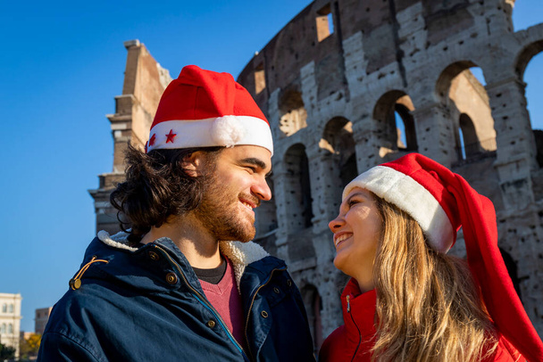 Римські свята на Різдво. Молода пара постає перед Колізеєм у червоних капелюхах Санта Клауса. Молоді люди дивляться одне одному в очі і посміхаються. Почуття щастя і любові. - Фото, зображення