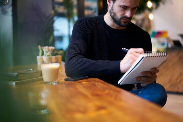 Crop concentrated bearded ethnic male in casual wear sitting at wooden table and taking notes in copybook while working remotely - Valokuva, kuva