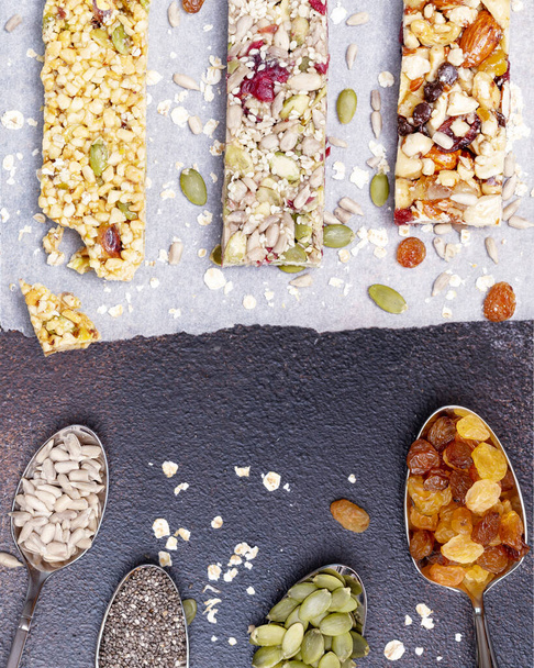 Different kind of granola cereal bars with nuts, seeds, oats, berries, dry fruits and spoons with ingredients on a dark brown background. Protein muesli bars. Granola superfood. Energy healthy snack - Photo, image