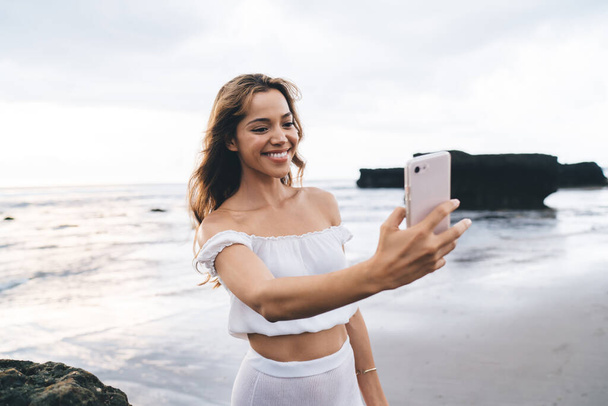 Young happy female in white summer wear standing on sandy seashore and photographing with mobile phone against sea and rocks - Φωτογραφία, εικόνα