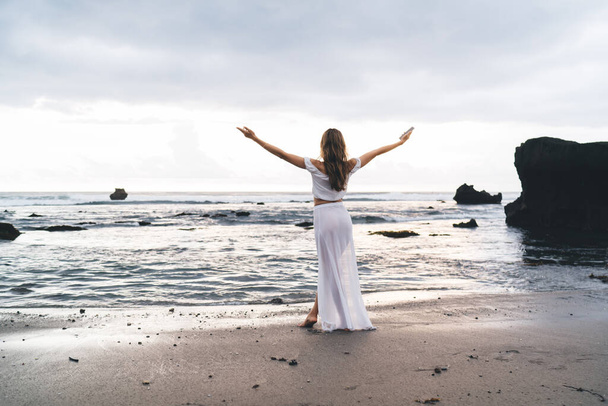 Back view of full length faceless slim barefoot female tourist in white dress standing on sandy beach with raised arms - Φωτογραφία, εικόνα