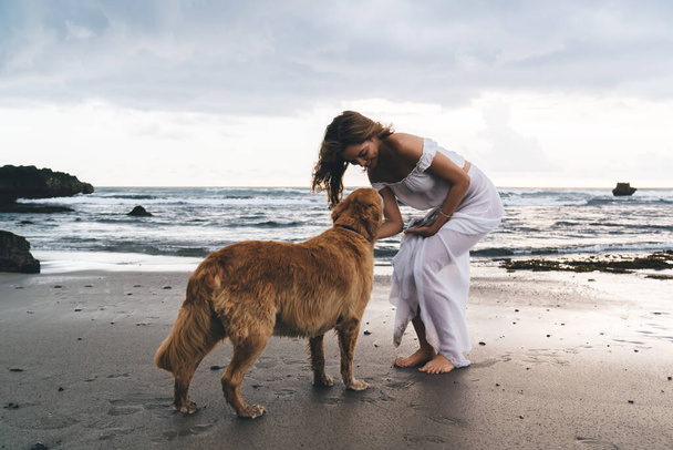 Full length of happy female in elegant wear standing on sandy seashore and caressing purebred Golden Retriever dog during stroll - Foto, afbeelding