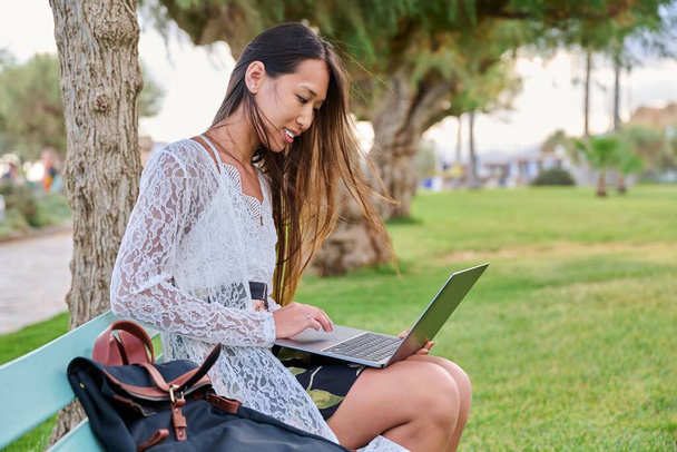 Beautiful young Asian woman using laptop sitting on an outdoor bench - Foto, Bild