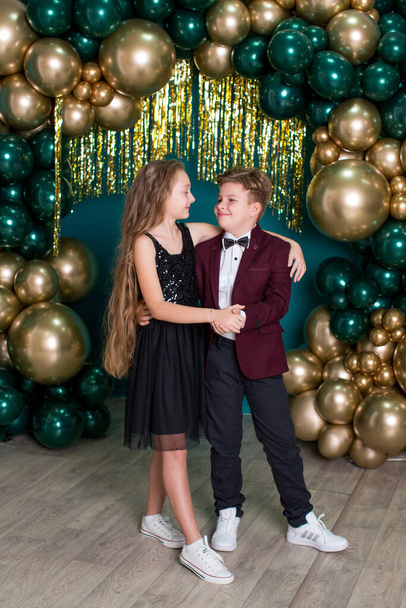 A girl with long hair in a black dress and a boy in a burgundy jacket and black trousers are hugging each other in an arch of gold and green balloons on a green background - Foto, imagen