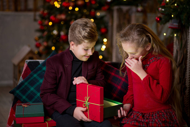 Niños: una niña con un vestido rojo y un niño con una chaqueta de color burdeos se sientan sobre el fondo de los árboles de Navidad. Abrir la caja de regalo - emite una luz brillante - Foto, Imagen