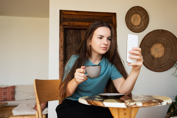 Красивая молодая женщина в повседневной рубашке сидит за столом в кафе и проверяет посыльного при использовании смартфона - Фото, изображение
