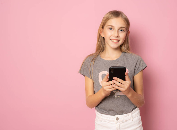 Smiling little girl in casual summer t-shirt using mobile phone standing isolated over pink background. - Photo, Image