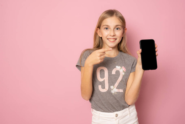 Улыбающаяся маленькая девочка в обычной летней футболке показывает экран мобильного телефона, стоящий изолированно на розовом фоне. - Фото, изображение