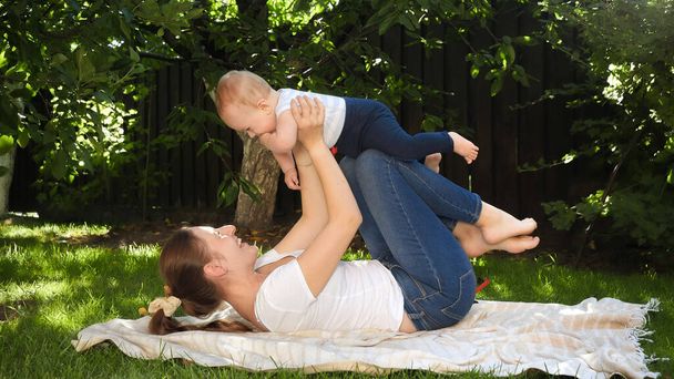 Madre sonriente levantándose y meciendo a su pequeño hijo en el jardín. Crianza, familia, desarrollo infantil y diversión al aire libre en la naturaleza. - Foto, Imagen