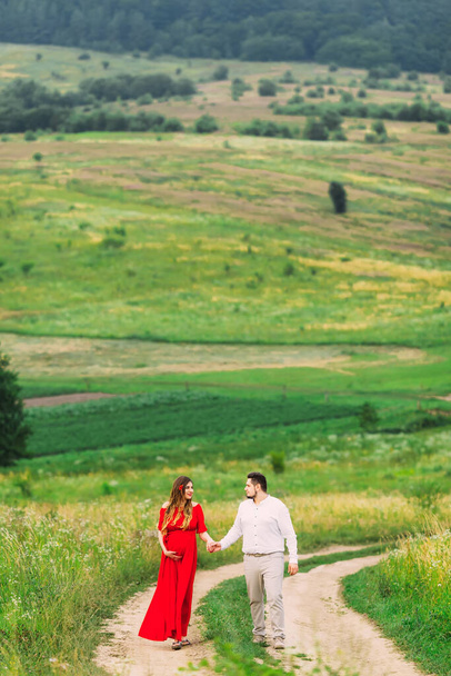 femme enceinte en robe rouge. les futurs parents se tiennent la main et marchent dans la prairie. - Photo, image