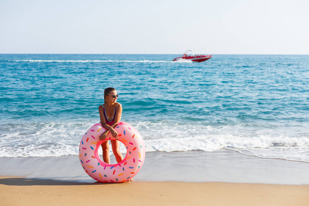 Привлекательная молодая женщина в ярком купальнике позирует на пляже с надувным кольцом. Красивая блондинка с длинными волосами отдыхает у океана. Концепция спортивной модели, купальники - Фото, изображение