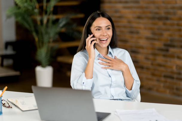 Phone call concept. Positive successful caucasian brunette woman, IT specialist, top manager, sit at work desk, having a nice cell phone conversation during work time, smile friendly, looks away - Фото, изображение