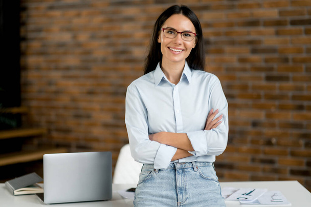 Portrait of successful intelligent pretty caucasian businesswoman, broker, real estate agent, stand near work desk in office, in formal stylish wear and glasses, arms crossed, looks at camera, smiles - Photo, Image