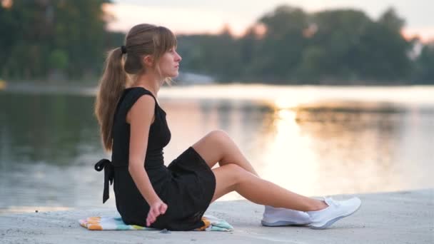 Donna sola seduta sul lato del lago la sera calda. Solitudine e concetto di relax - Filmati, video