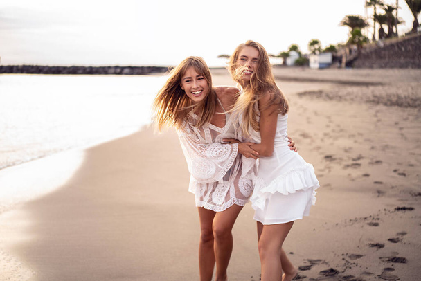 Two young blonde caucasian women walking on the beach, playing and laughing. Female friends having fun on the sea shore. Real people emotions. Happy lifestyle. Travel. - Photo, Image