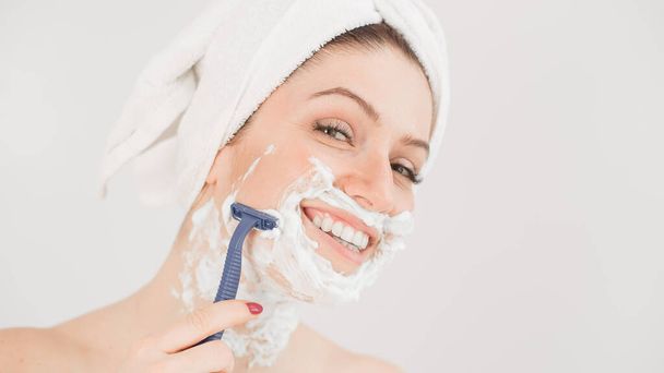Cheerful caucasian woman with a towel on her head and shaving foam on her face holds a razor on a white background - Fotografie, Obrázek