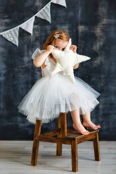 A cute little girl in an elegant dress is having fun, playing and hiding behind a textile star on a dark gray background with a garland of flags. Studio portrait of a child. - Foto, Imagem