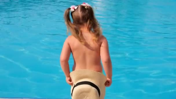 grappig klein meisje 3 jaar oud danst en verstopt zich achter een zonnehoed op de achtergrond van blauw water van het zwembad of de zee, achteraanzicht. Het concept van zomervakantie, zonnebrandcrème, reizen. selectieve focus op - Video
