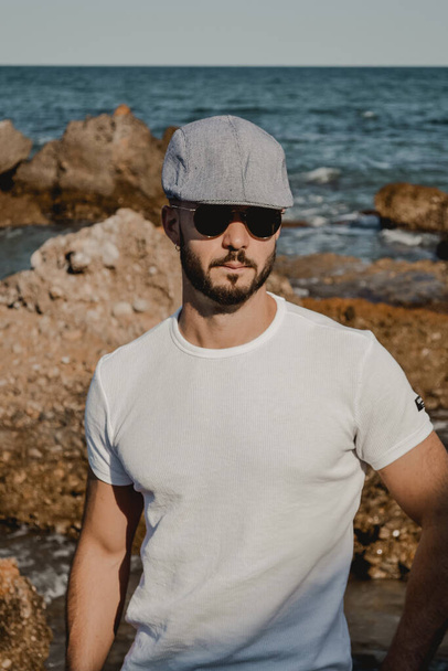 A vertical shot of a cool man wearing an ivy cap and sunglasses near the sea - Foto, Imagem