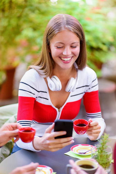 Mujer joven feliz sonriendo en deleite mientras usa su teléfono inteligente mientras se relaja en una mesa tomando una taza de café con amigos en un restaurante al aire libre - Foto, Imagen