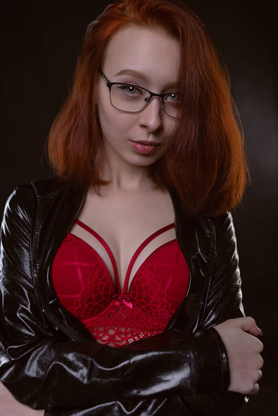 young sexy female model in a black latex raincoat and red beautiful underwear, portrait on a black background - Фото, изображение