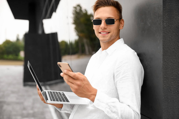 Młody człowiek w okularach przeciwsłonecznych z laptopem za pomocą telefonu komórkowego na zewnątrz - Zdjęcie, obraz