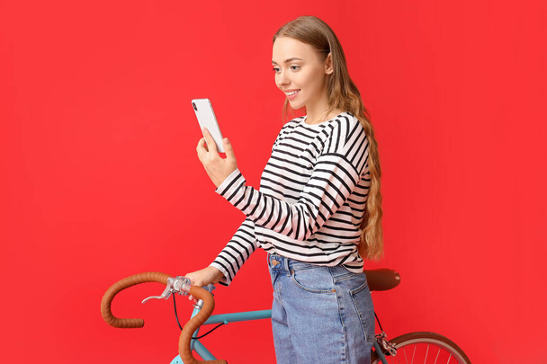 Όμορφη έφηβη με ποδήλατο χρησιμοποιώντας το κινητό τηλέφωνο σε κόκκινο φόντο - Φωτογραφία, εικόνα