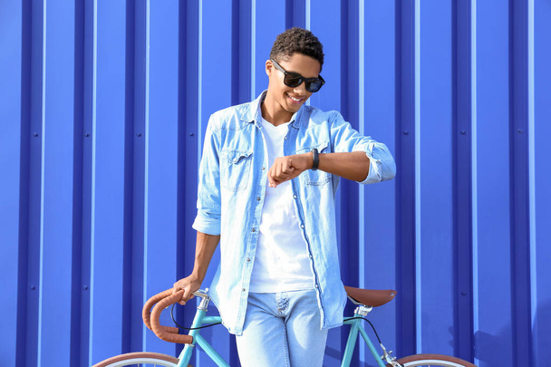 Adolescente afroamericano con bicicleta mirando un reloj de pulsera cerca de la cerca azul - Foto, Imagen
