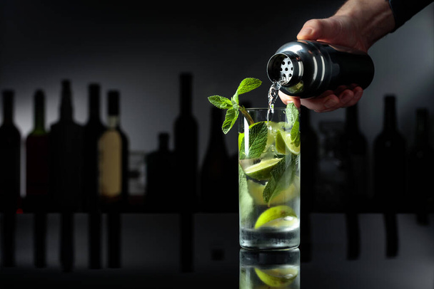 Cocktail "Mojito" auf schwarzem reflektierendem Hintergrund. Der Barkeeper gießt einen Cocktail aus einem Shaker in ein Glas mit Eis. - Foto, Bild