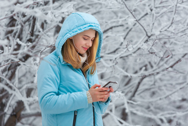 スマートフォンでオンラインで動画を見たり、雪の冬の公園のフードを歩いている間にヘッドフォンで音楽を聴いたりするかわいい笑顔の若い女性の写真 - 写真・画像