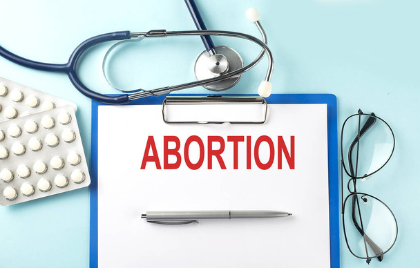 Papel con texto ABORTION sobre fondo azul con estetoscopio y pastillas - Foto, imagen