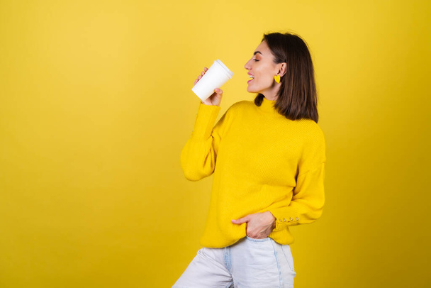 Jovem mulher em um suéter aconchegante amarelo com brilho labial rosa brilhante com uma xícara de papel de café aromático quente, looks sonhadores, sorriso bonito, calor irradiante - Foto, Imagem