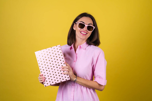 Mujer joven con estilo en un vestido rosa y gafas sobre un fondo amarillo con una caja de regalo en sus manos celebra la mejor fiesta, cumpleaños, Día de San Valentín - Foto, Imagen