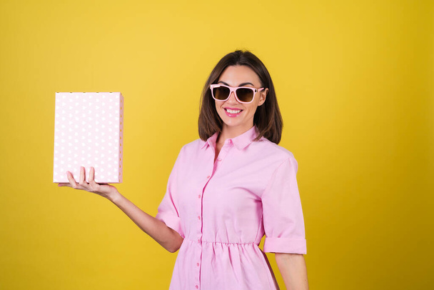 Elegante giovane donna in un abito rosa e occhiali su uno sfondo giallo con una scatola regalo in mano celebra la migliore vacanza, compleanno, San Valentino - Foto, immagini