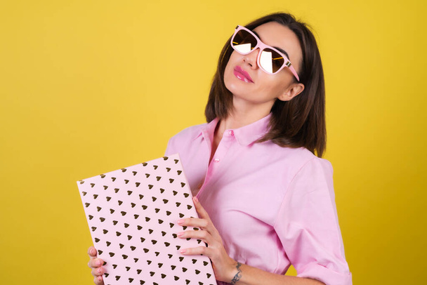 Elegante giovane donna in un abito rosa e occhiali su uno sfondo giallo con una scatola regalo in mano celebra la migliore vacanza, compleanno, San Valentino - Foto, immagini