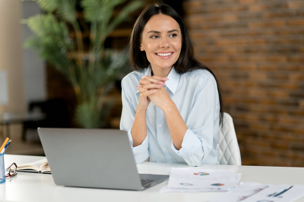Привлекательная кавказская брюнетка деловая женщина, наставница, офисный работник, финансовый консультант, сидит за столом в офисе, отворачивается, улыбается дружелюбно, отдыхает от работы - Фото, изображение