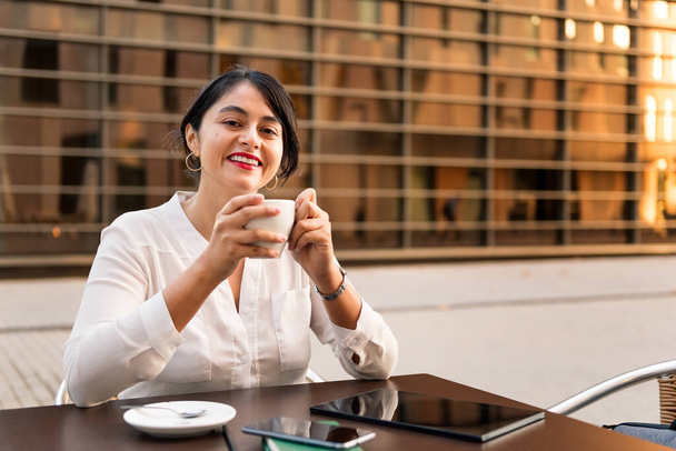 businesswoman smiling with a cup of coffee in her hands on the terrace of a coffee shop, concept of digital entrepreneur and urban lifestyle, copy space for text - Фото, изображение