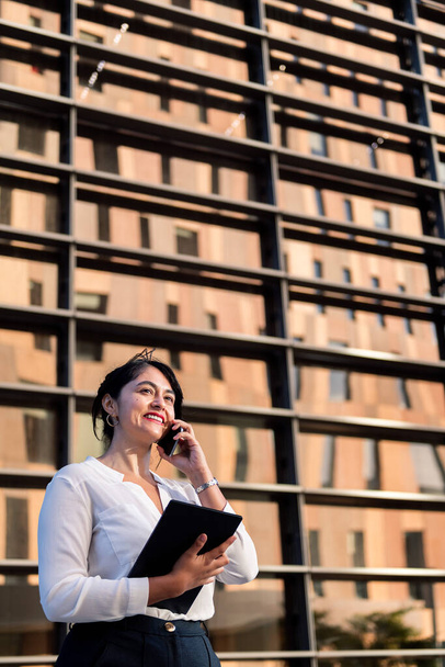 smiling businesswoman talking on the phone and using her tablet in front of an office building, concept of successful entrepreneur and urban lifestyle, copy space for text - Fotografie, Obrázek