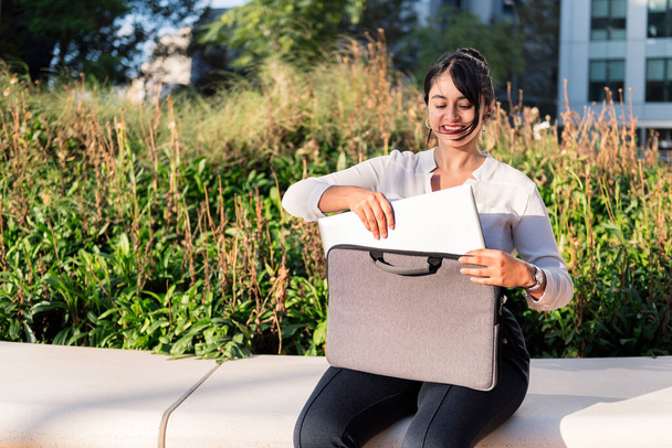 smiling businesswoman taking her laptop computer out of her briefcase sitting in a park, concept of digital entrepreneur and urban lifestyle, copy space for text - 写真・画像