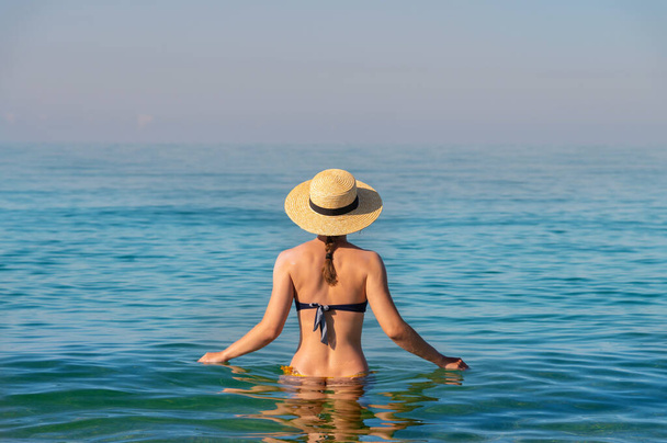 Back view of a slender caucasian young woman standing waist-deep in calm transparent sea water in a swimsuit and a straw hat - Fotografie, Obrázek